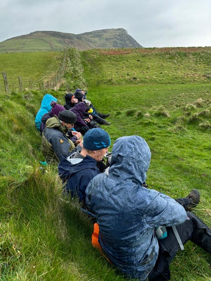 5.Porth Meudwy - Mynydd Anelog
28/3/24. The group tuck into lunch on the Wales Coast Path just below Mynydd Anelog. Mynydd Mawr, at the end of the peninsula,r in the background. Photo: Louise Fletcher-Brewer.
Keywords: Mar24 Thursday Annie Andrew Jean Norton